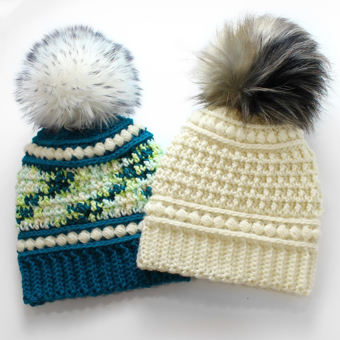 May include: Two handmade crochet beanies with faux fur pom-poms. One beanie is teal with white and green accents, the other is cream-colored. Both have a ribbed brim and decorative stitching.