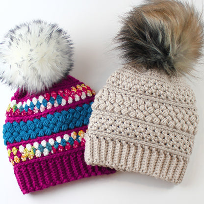 May include: Two crocheted hats with faux fur pom poms. One hat is a bright pink with colorful stripes and a white pom pom. The other hat is beige with a brown and black pom pom.