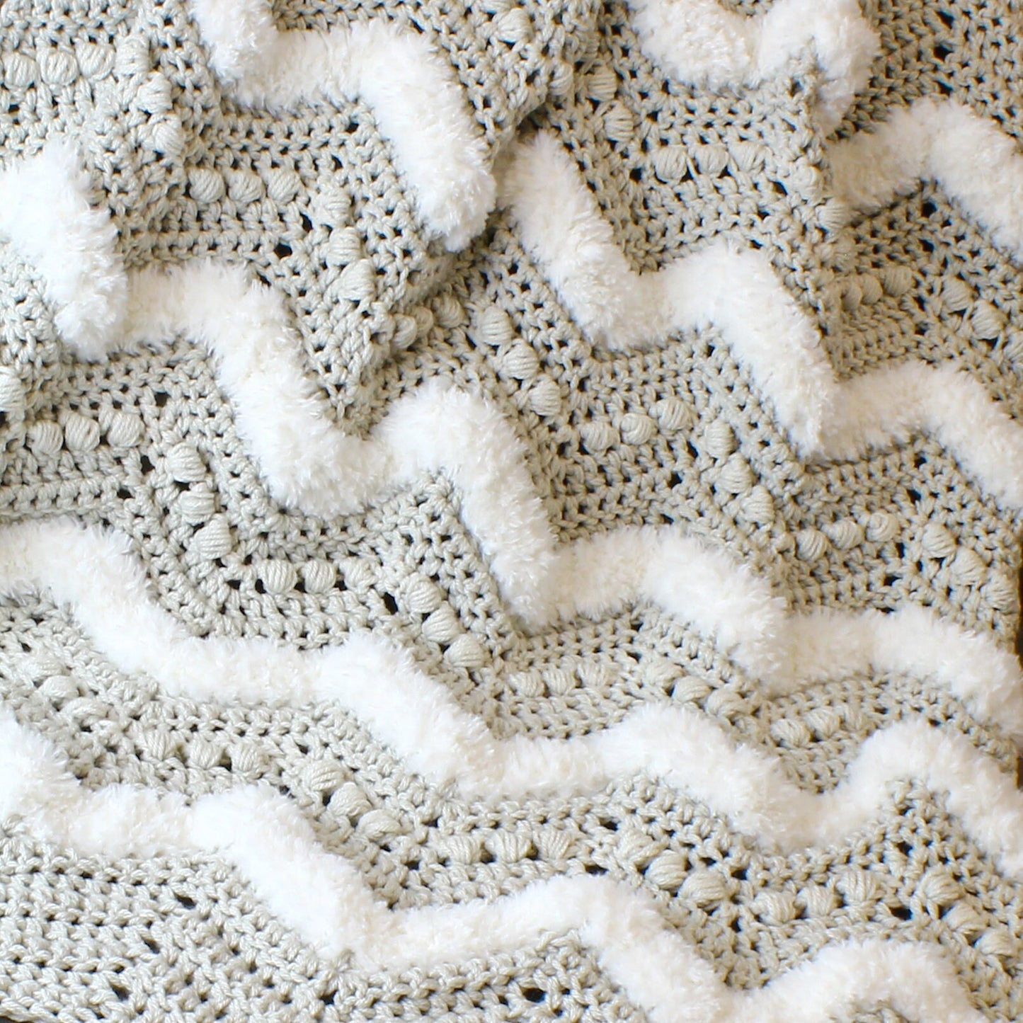 May include: A close-up of a crocheted blanket with a chevron pattern. The blanket is made with a light gray yarn and has a white fuzzy trim.