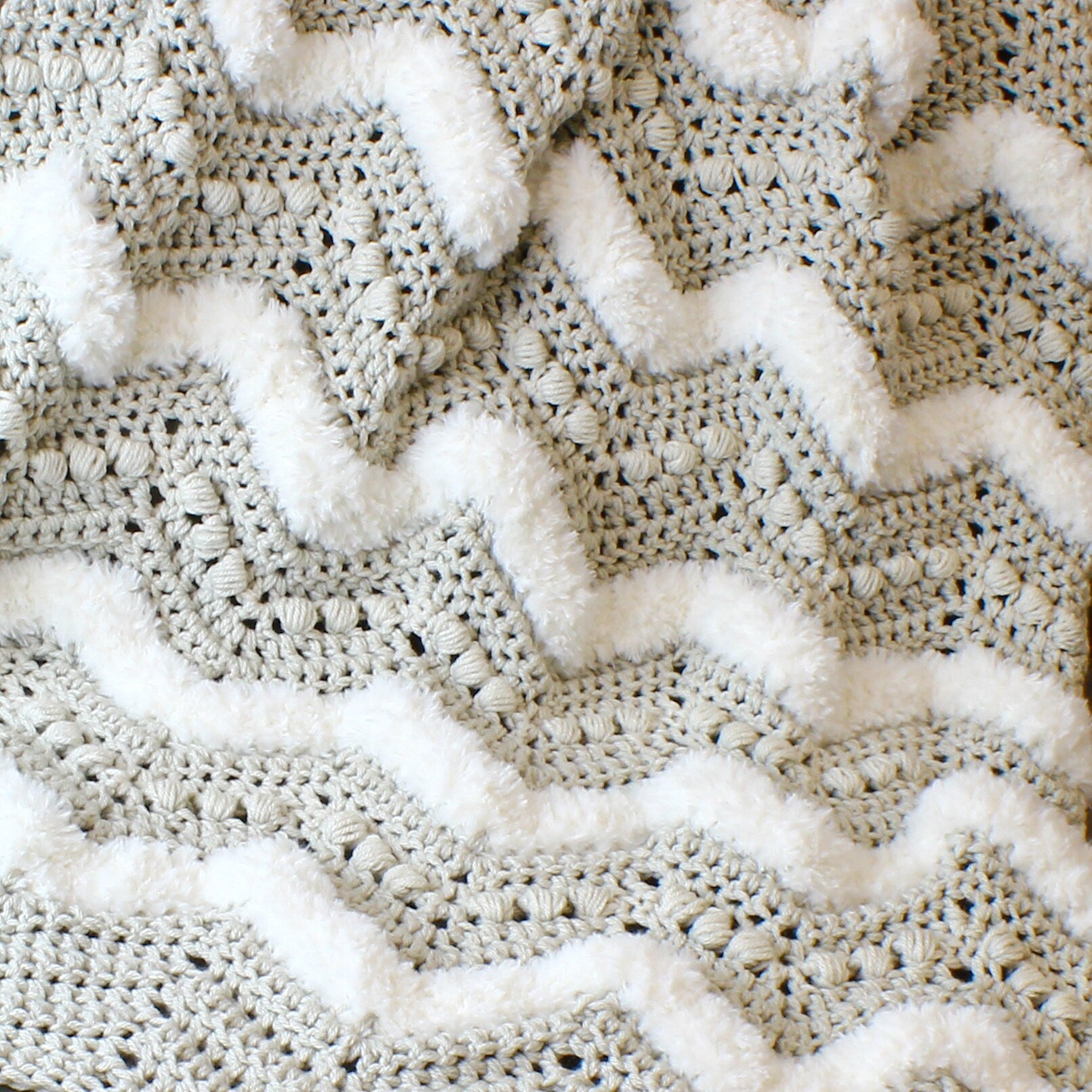 May include: A close-up of a crocheted blanket with a chevron pattern. The blanket is made with a light gray yarn and has a white fuzzy trim.