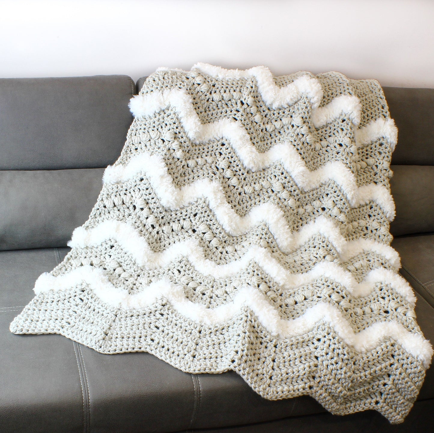 May include: A gray and white crocheted throw blanket with a chevron pattern and white pom-poms along the edges. The blanket is draped over a gray couch.