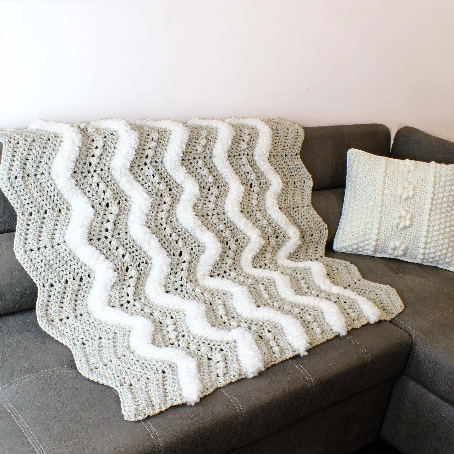 May include: A gray and white crocheted throw blanket with a wavy pattern and white pom-poms along the edges. The blanket is draped over a gray couch.