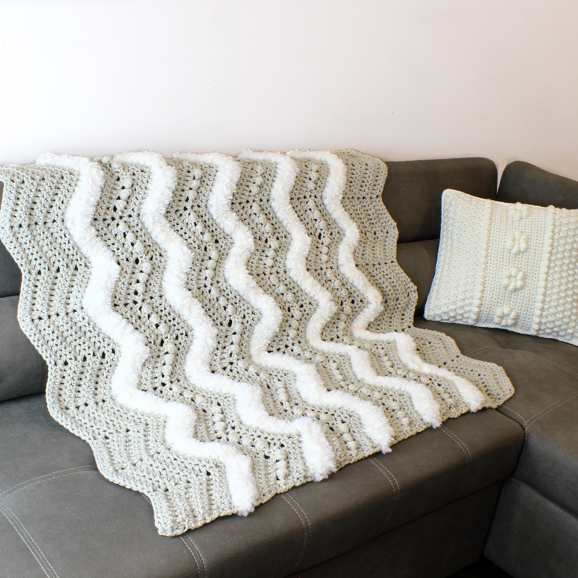May include: A gray and white crocheted throw blanket with a wavy pattern and white pom-poms along the edges. The blanket is draped over a gray couch.
