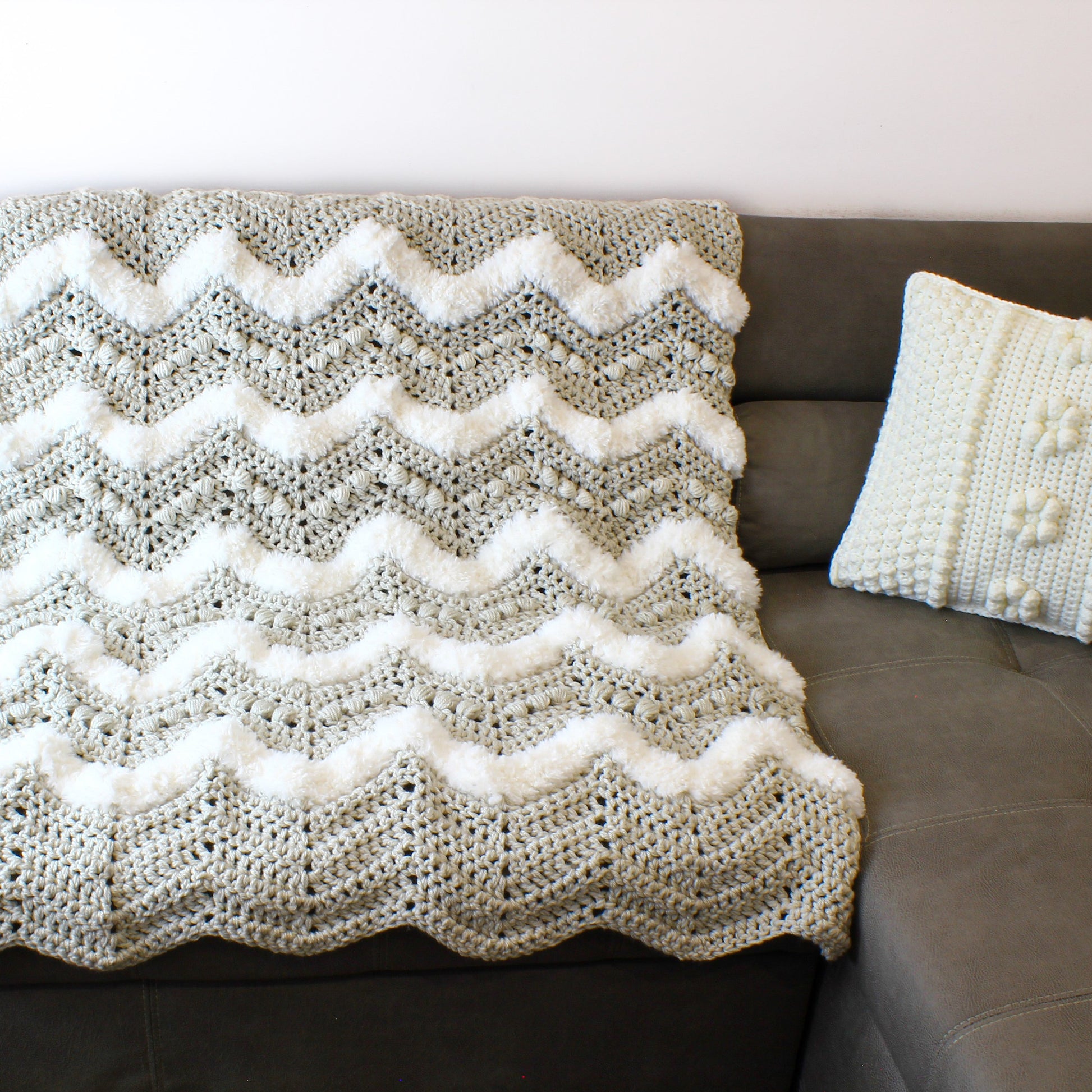 May include: A gray and white crocheted throw blanket with a chevron pattern. The blanket is draped over a brown couch.