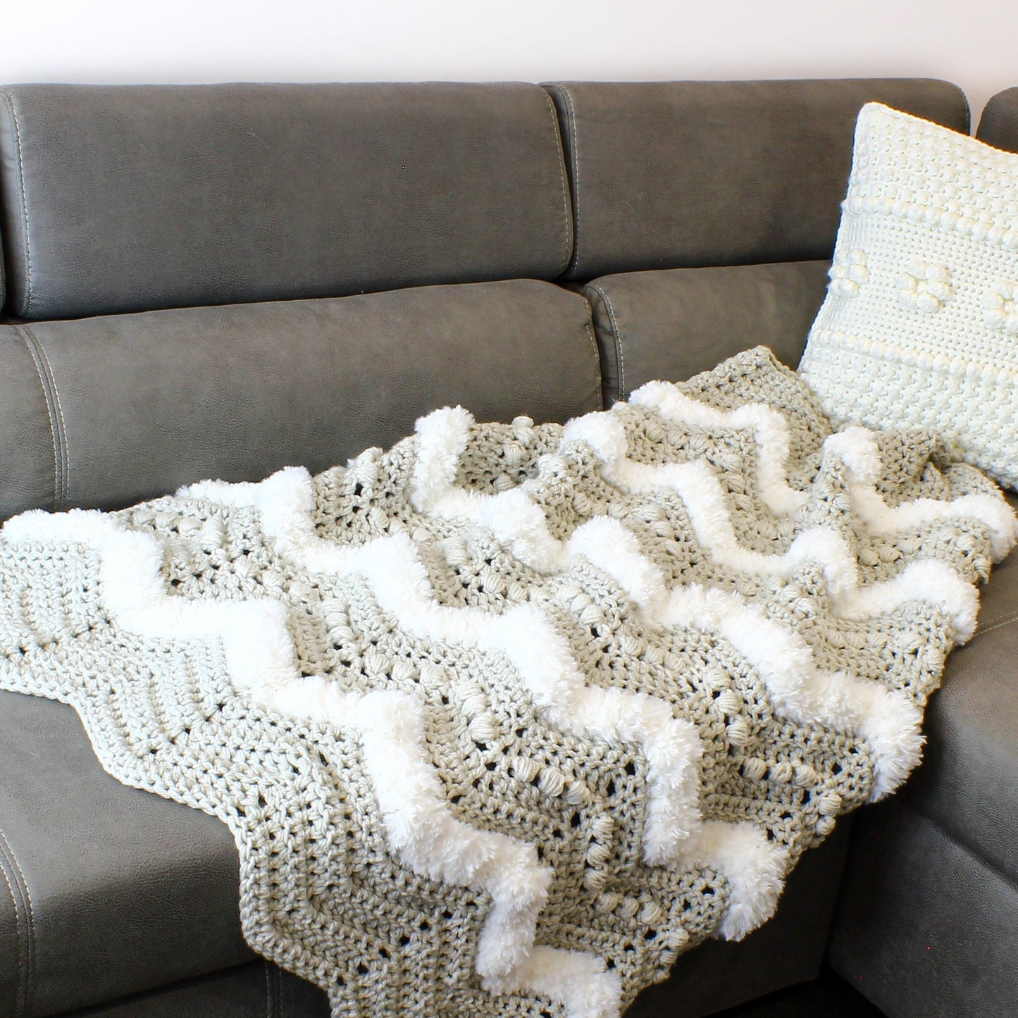 May include: A crocheted throw blanket with a chevron pattern and white pom-poms. The blanket is draped over a gray couch.