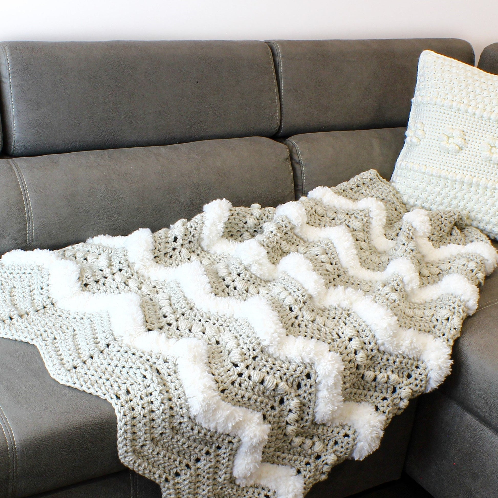May include: A crocheted throw blanket with a chevron pattern and white pom-poms. The blanket is draped over a gray couch.