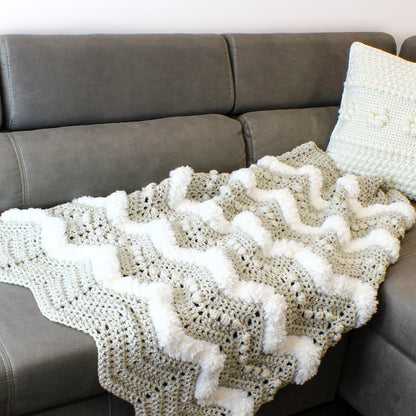 May include: A crocheted throw blanket with a chevron pattern and white pom-poms. The blanket is draped over a gray couch.