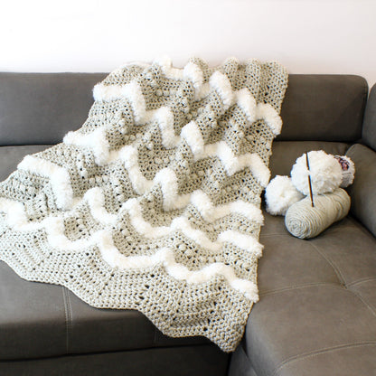 May include: A crocheted throw blanket with a chevron pattern in sage green and white. The blanket has a fluffy texture and is made with a chunky yarn.