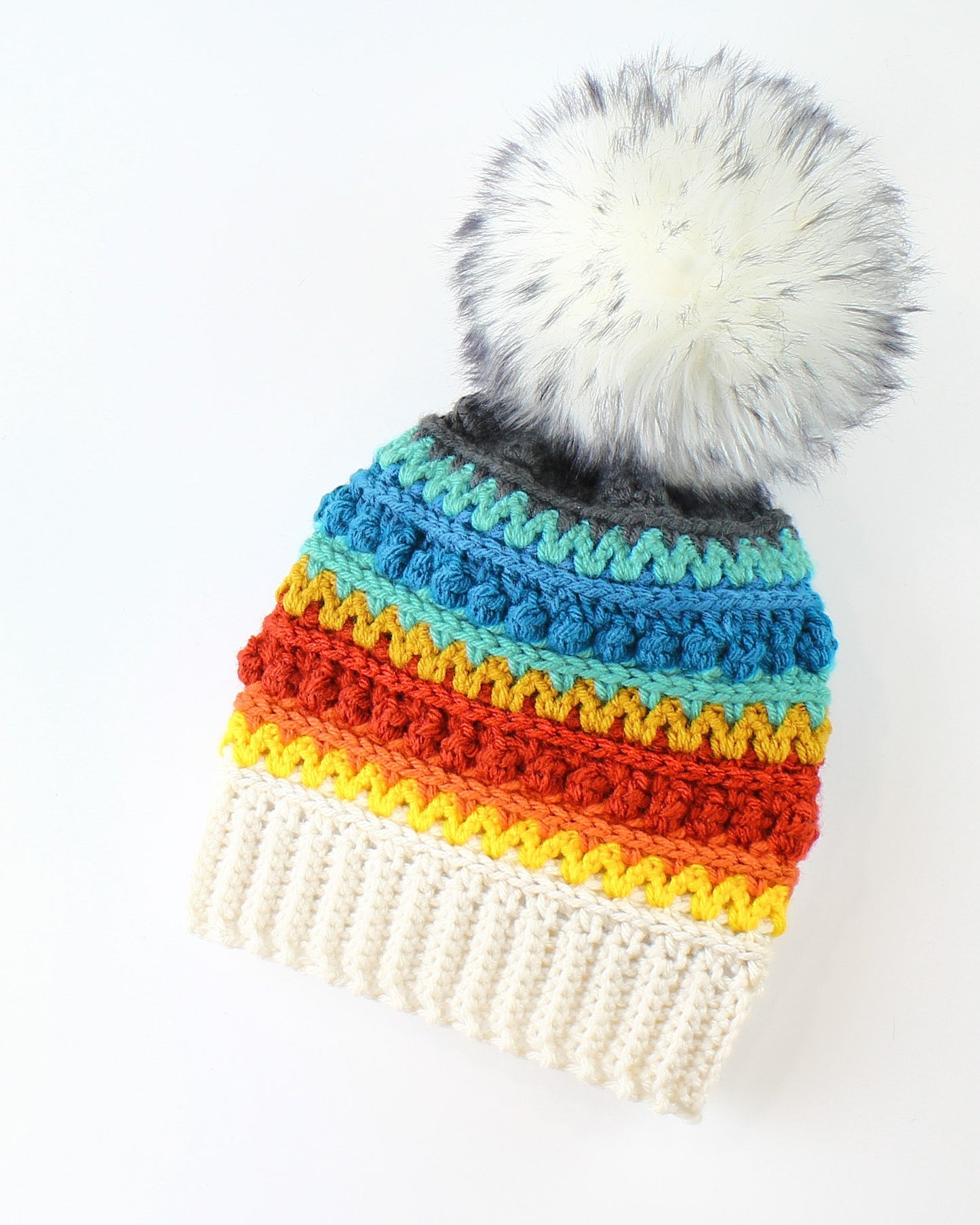 May include: A colorful crocheted beanie with a white faux fur pom pom. The beanie features stripes of red, orange, yellow, green, blue, and gray.