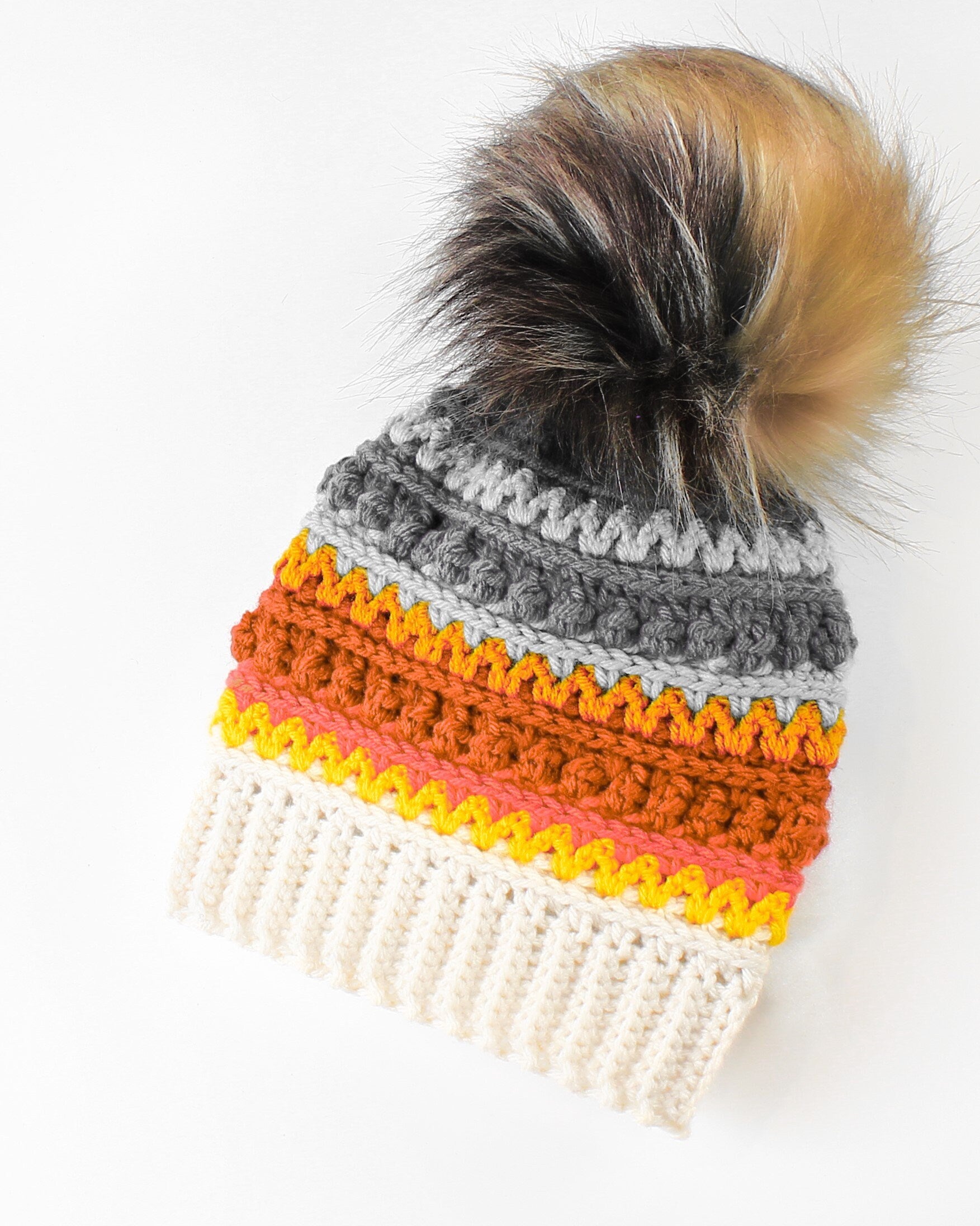 May include: A crocheted beanie with a gray, orange, yellow, and white striped pattern. The beanie has a fluffy black and brown pom pom on top.