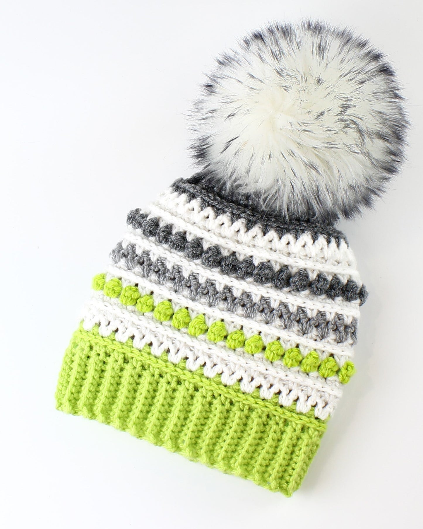 May include: A green, white, and gray striped crocheted beanie with a white faux fur pom pom.