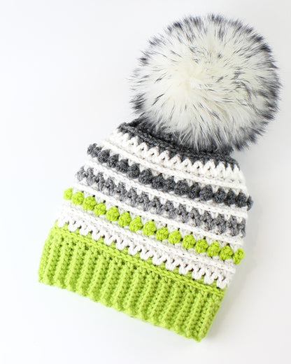 May include: A green, white, and gray striped crocheted beanie with a white faux fur pom pom.