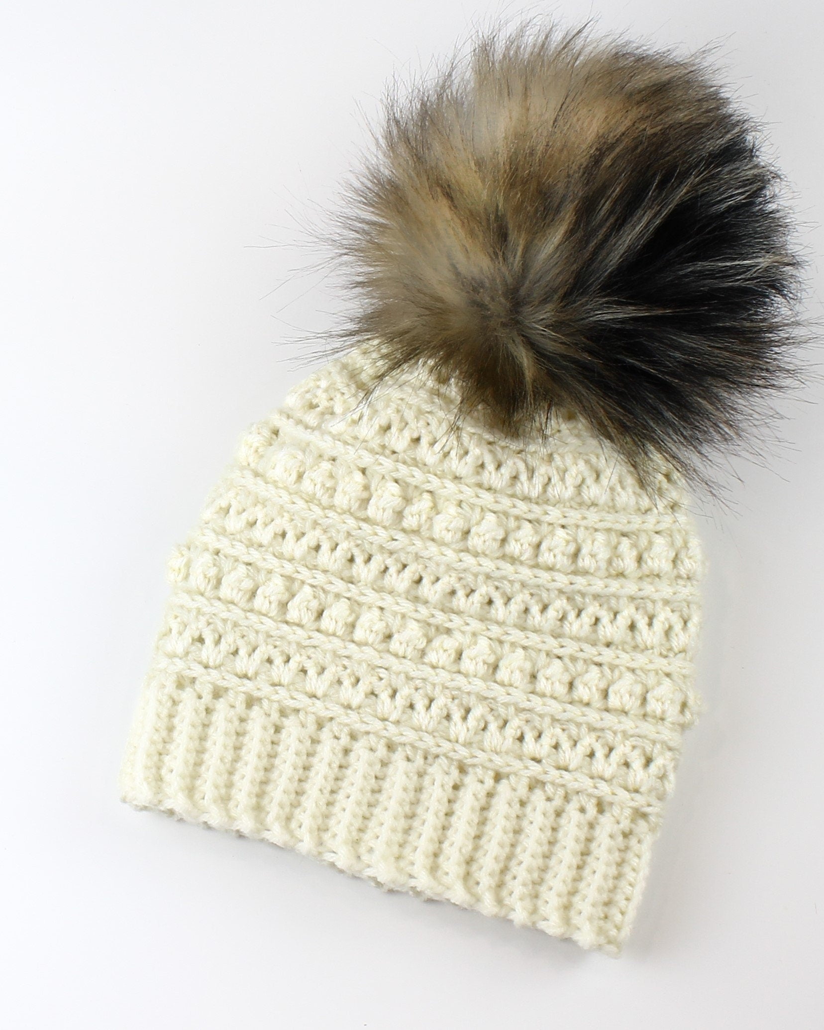 May include: A white crocheted beanie with a brown and black faux fur pom pom.