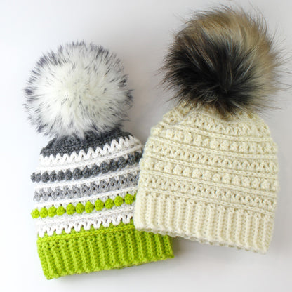 May include: Two crocheted hats with faux fur pom poms. One hat is white with a grey and green striped pattern. The other hat is a solid cream color.