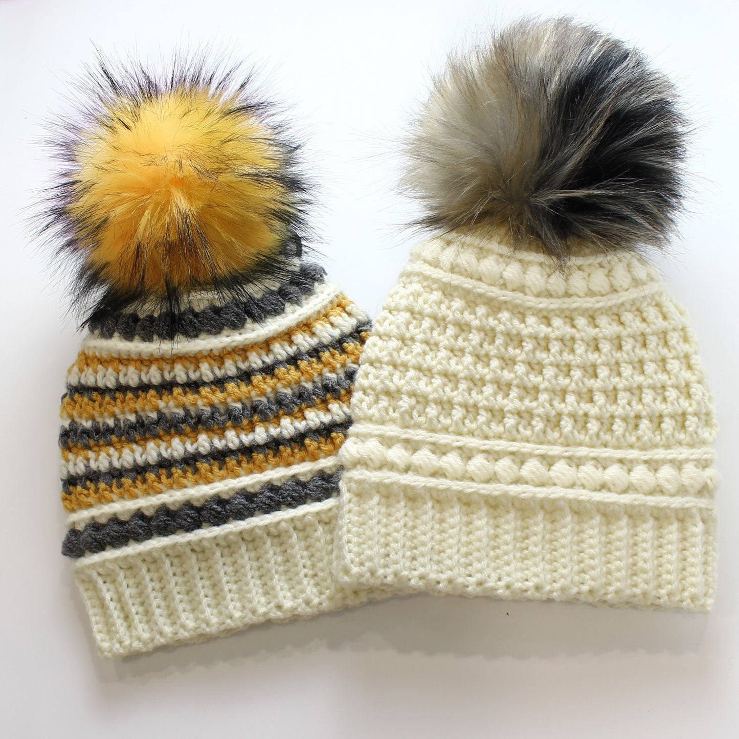 May include: Two handmade crochet beanies with faux fur pom-poms. One beanie is cream-colored with a textured pattern, and the other is striped with cream, gray, and mustard yellow. Both have ribbed brims.