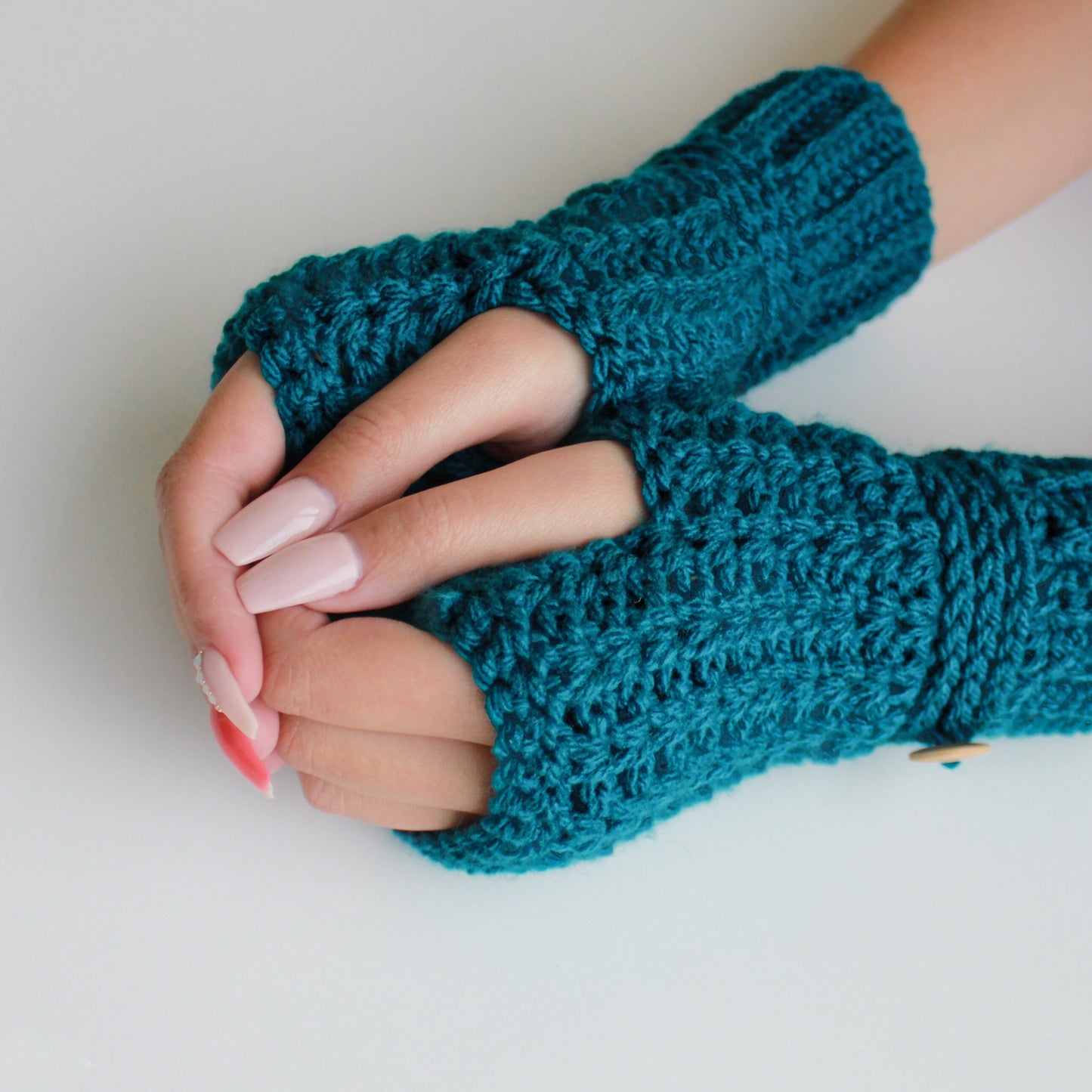 May include: Teal blue fingerless gloves with a crocheted texture. The gloves have a button detail on the wrist.