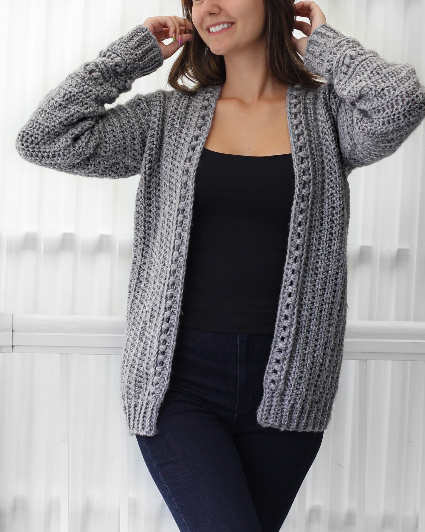 May include: A gray crocheted cardigan with a V-neck and long sleeves. The cardigan has a textured stitch pattern and a delicate openwork design along the front edges.