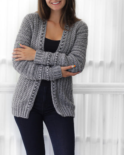 May include: A gray crocheted cardigan with a V-neck and long sleeves. The cardigan has a textured stitch pattern and a lace trim along the front and cuffs.