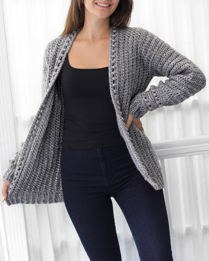 May include: A gray crocheted cardigan with a ribbed texture and a crocheted trim around the neckline and cuffs. The cardigan is open in the front and has long sleeves.