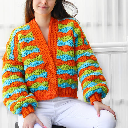 May include: A crocheted cardigan with a colorful striped pattern in orange, green, and blue. The cardigan has a button-up front and long sleeves with a slightly puffed look.