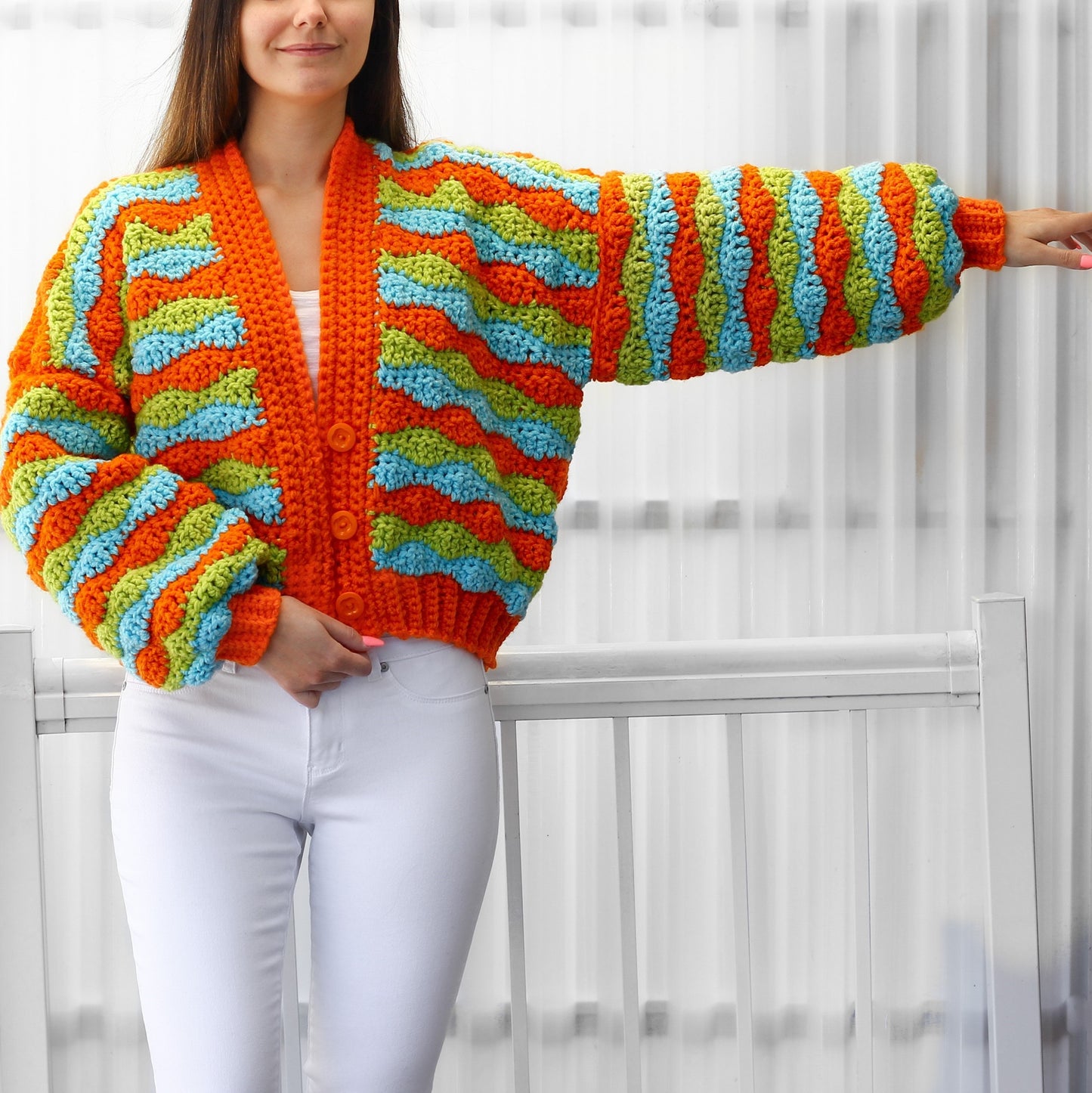 May include: A crocheted cardigan with a wavy pattern in orange, green, and blue. The cardigan has a button-up front and long sleeves.
