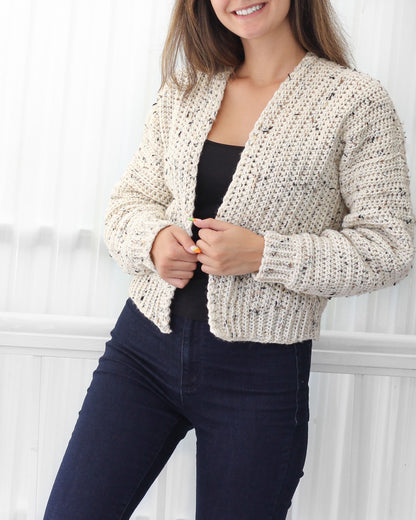 May include: A cream-colored crocheted cardigan with a speckled pattern. The cardigan has a V-neck and long sleeves. The model is wearing the cardigan with a black tank top and dark blue jeans.