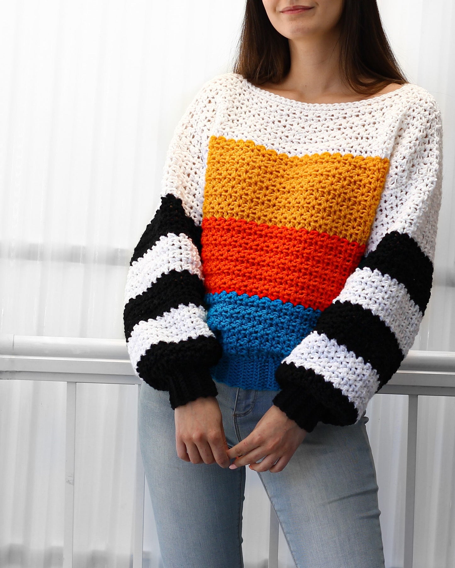 May include: A colorful crocheted sweater with a striped pattern on the sleeves and a horizontal stripe pattern on the body. The sweater is white, yellow, orange, blue, and black.