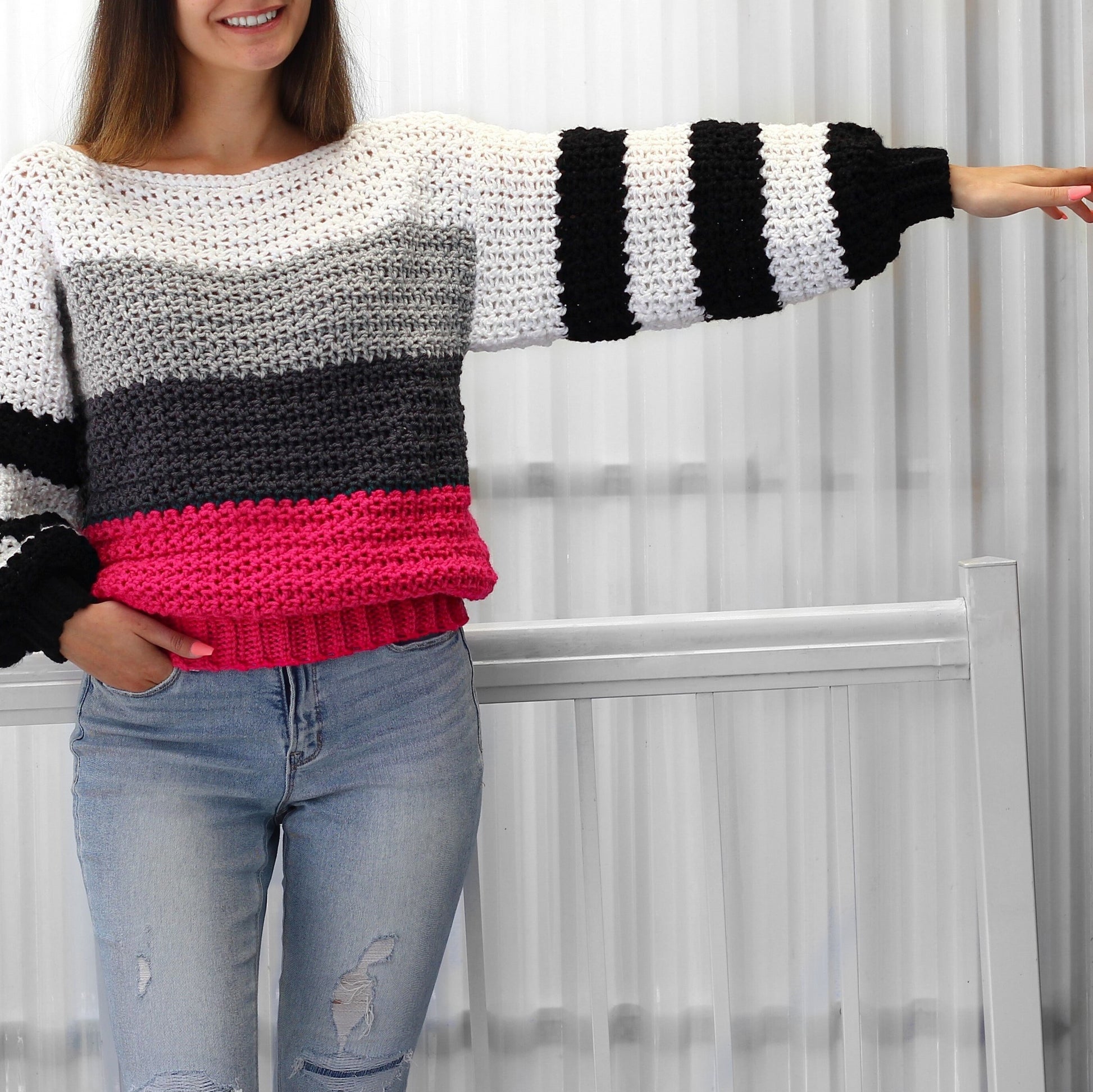May include: A striped crochet sweater with a white, gray, black, and pink color scheme. The sweater has long sleeves with a wide, striped cuff. The sweater is made with a chunky yarn and has a relaxed fit.