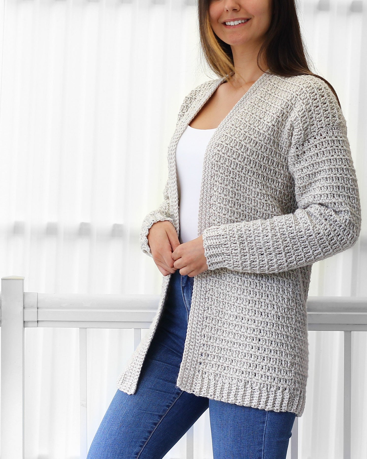 May include: A light gray crocheted cardigan with a V-neck and long sleeves. The cardigan is open in the front and is being worn over a white tank top and blue jeans.