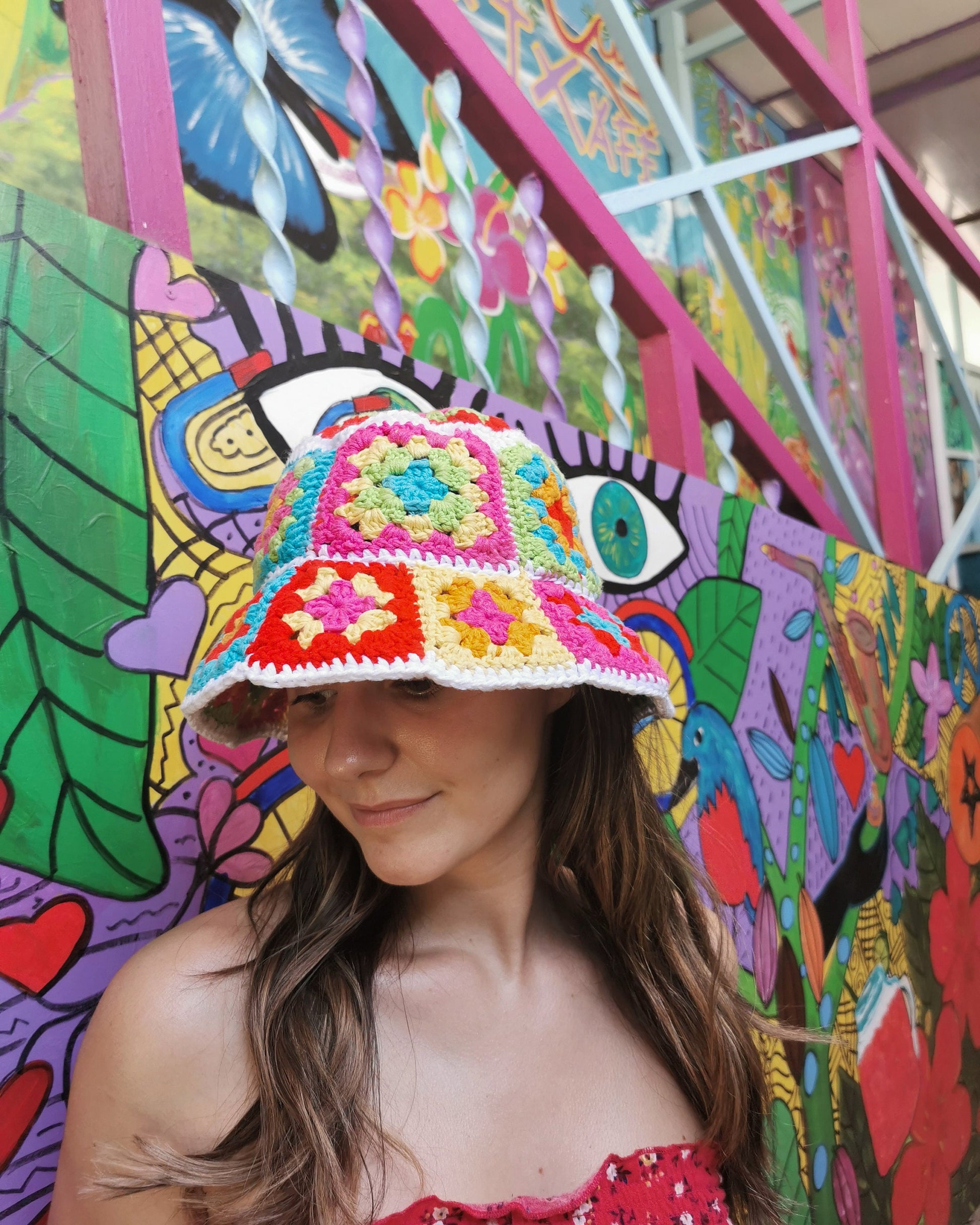 May include: A woman wearing a colorful crocheted bucket hat with a square pattern. The hat is made with bright pink, yellow, blue, and green yarn. The woman is standing in front of a colorful wall with a mural.
