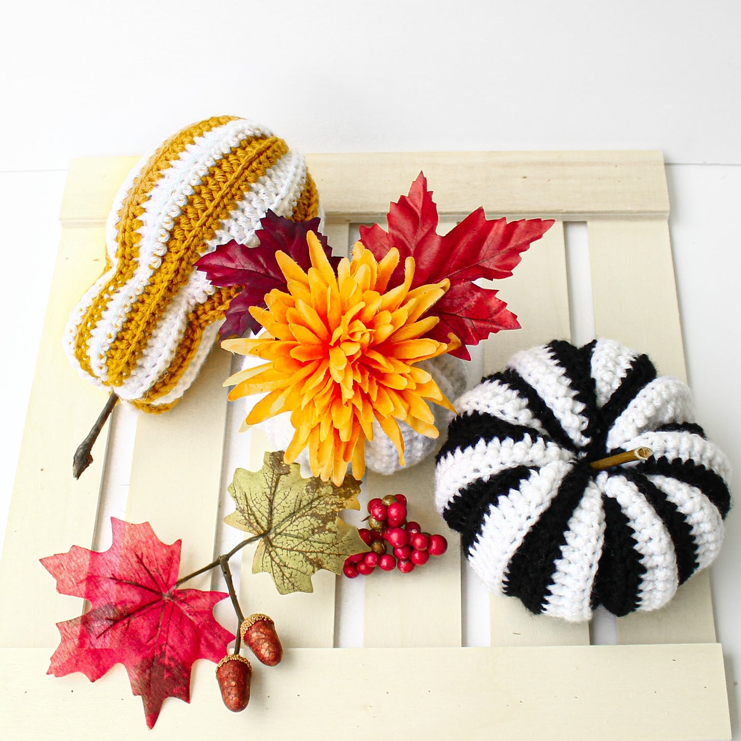 May include: Three crocheted pumpkins, one yellow and white striped, one black and white striped, and one white with a yellow flower on top. The pumpkins are on a white wooden surface with red berries and fall leaves.