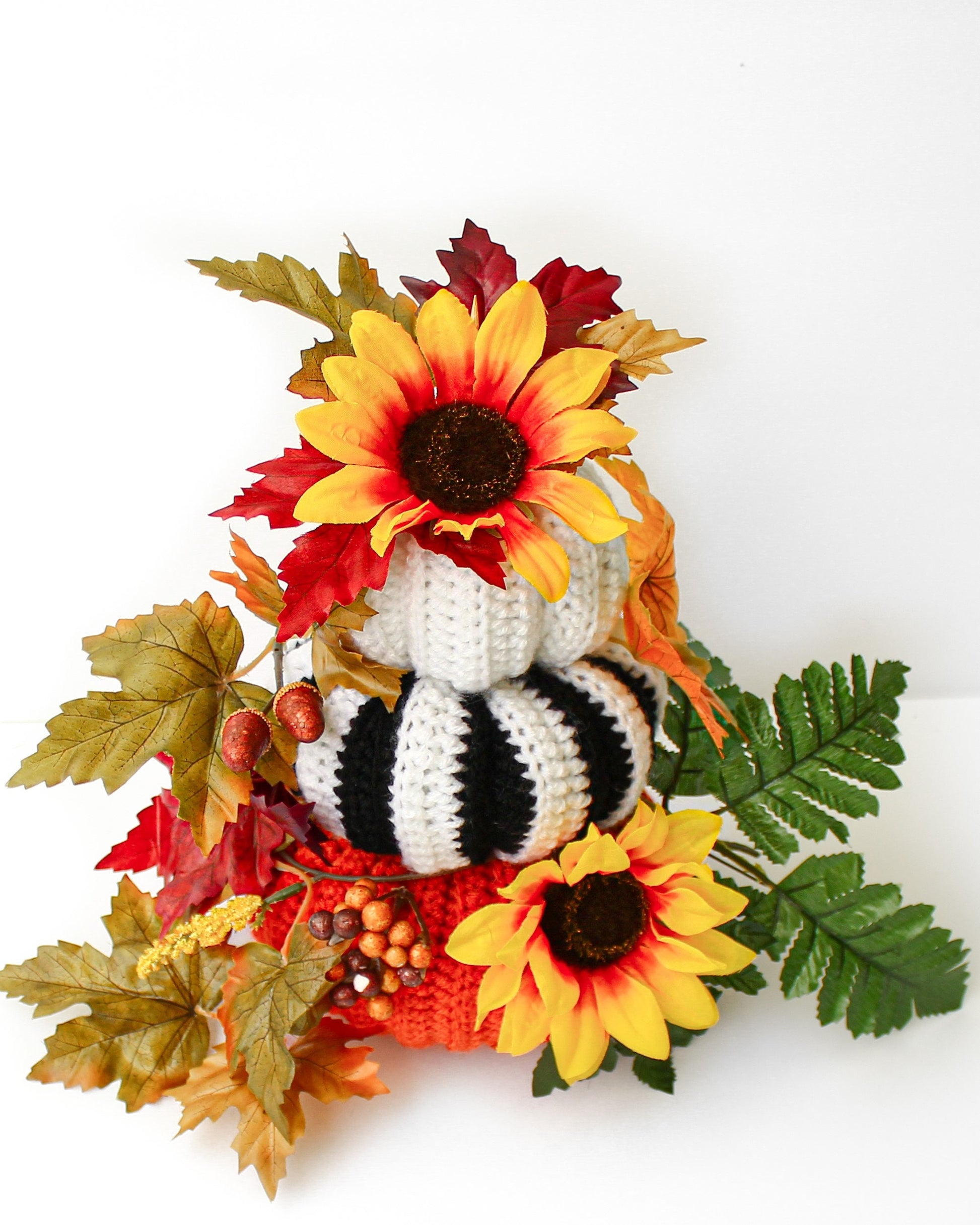 May include: A stack of three crocheted pumpkins decorated with fall foliage and sunflowers. The pumpkins are white, black and white striped, and orange.