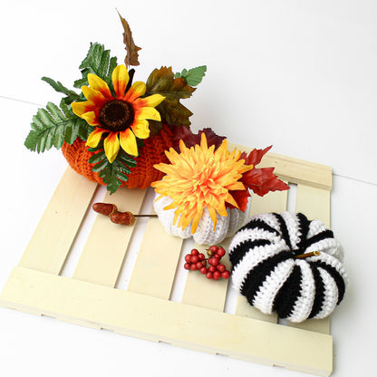 May include: Three crocheted pumpkins on a wooden crate. The pumpkins are decorated with fall-themed flowers and berries. The pumpkins are orange, white, and black and white striped.