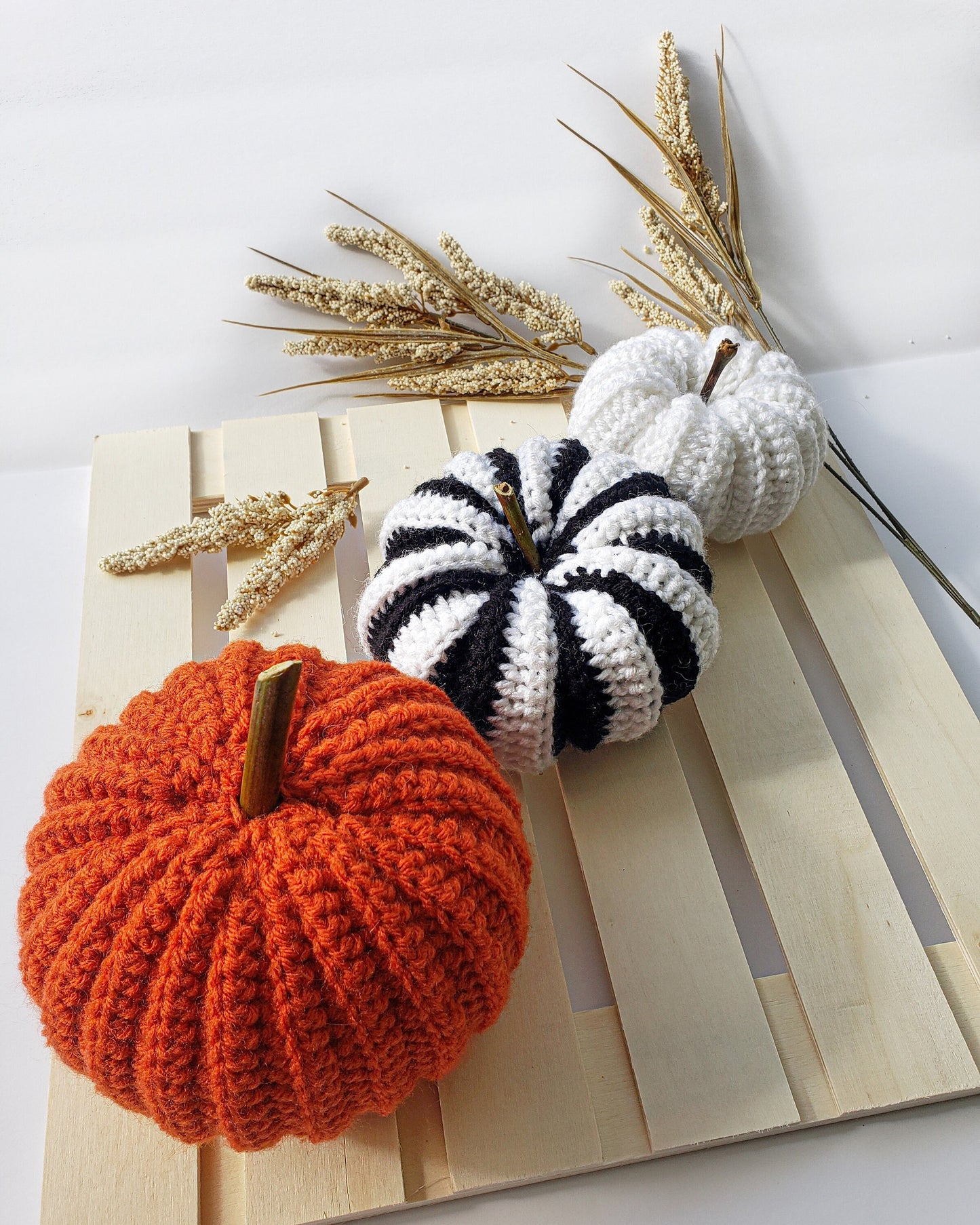 May include: Three crocheted pumpkins on a wooden pallet. One pumpkin is orange, one is white, and one is black and white striped.