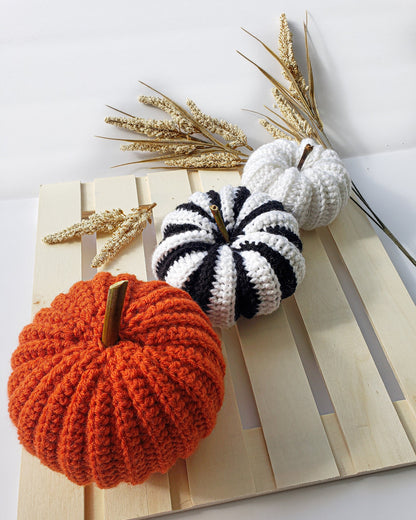 May include: Three crocheted pumpkins on a wooden pallet. One pumpkin is orange, one is white, and one is black and white striped.