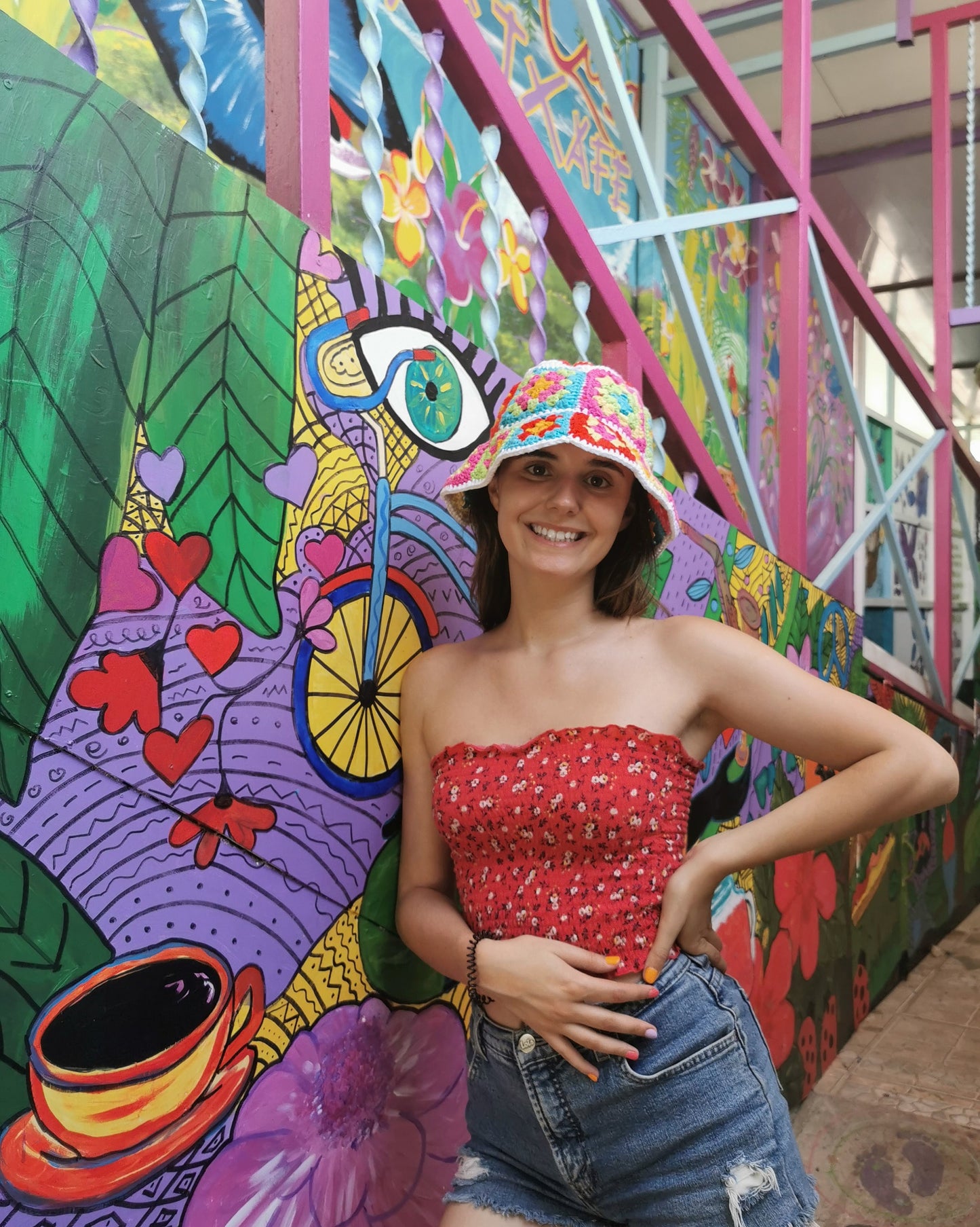May include: A woman wearing a colorful crocheted hat, a red floral print strapless top, and blue denim shorts. She is standing in front of a colorful mural with a bicycle, hearts, and flowers.