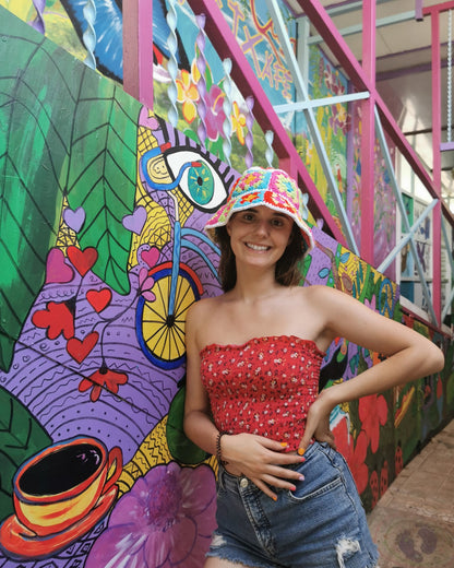 May include: A woman wearing a colorful crocheted hat, a red floral print strapless top, and blue denim shorts. She is standing in front of a colorful mural with a bicycle, hearts, and flowers.