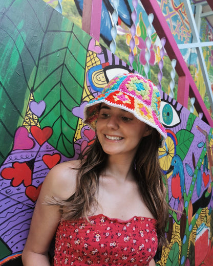 May include: A woman wearing a colorful crocheted bucket hat with a white brim. The hat has a geometric pattern with squares of pink, yellow, blue, green, and red. The woman is smiling and looking to the side. She is standing in front of a colorful wall with a mural of hearts, leaves, and other abstract designs.
