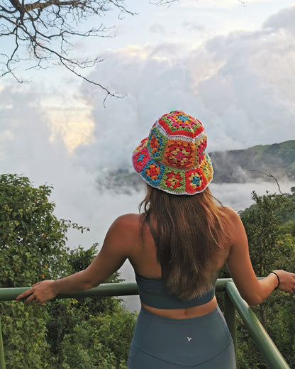 May include: A woman wearing a colorful crocheted bucket hat with a white background and a green, blue, pink, orange, and yellow pattern. She is standing on a platform overlooking a foggy landscape.