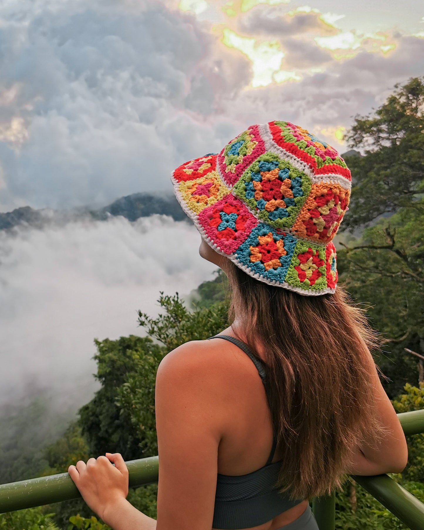 May include: A woman wearing a colorful crochet bucket hat with a white background and a variety of colors including red, orange, yellow, green, blue, and purple. The hat is worn on her head and she is looking out at a view of a mountain range with clouds in the distance.