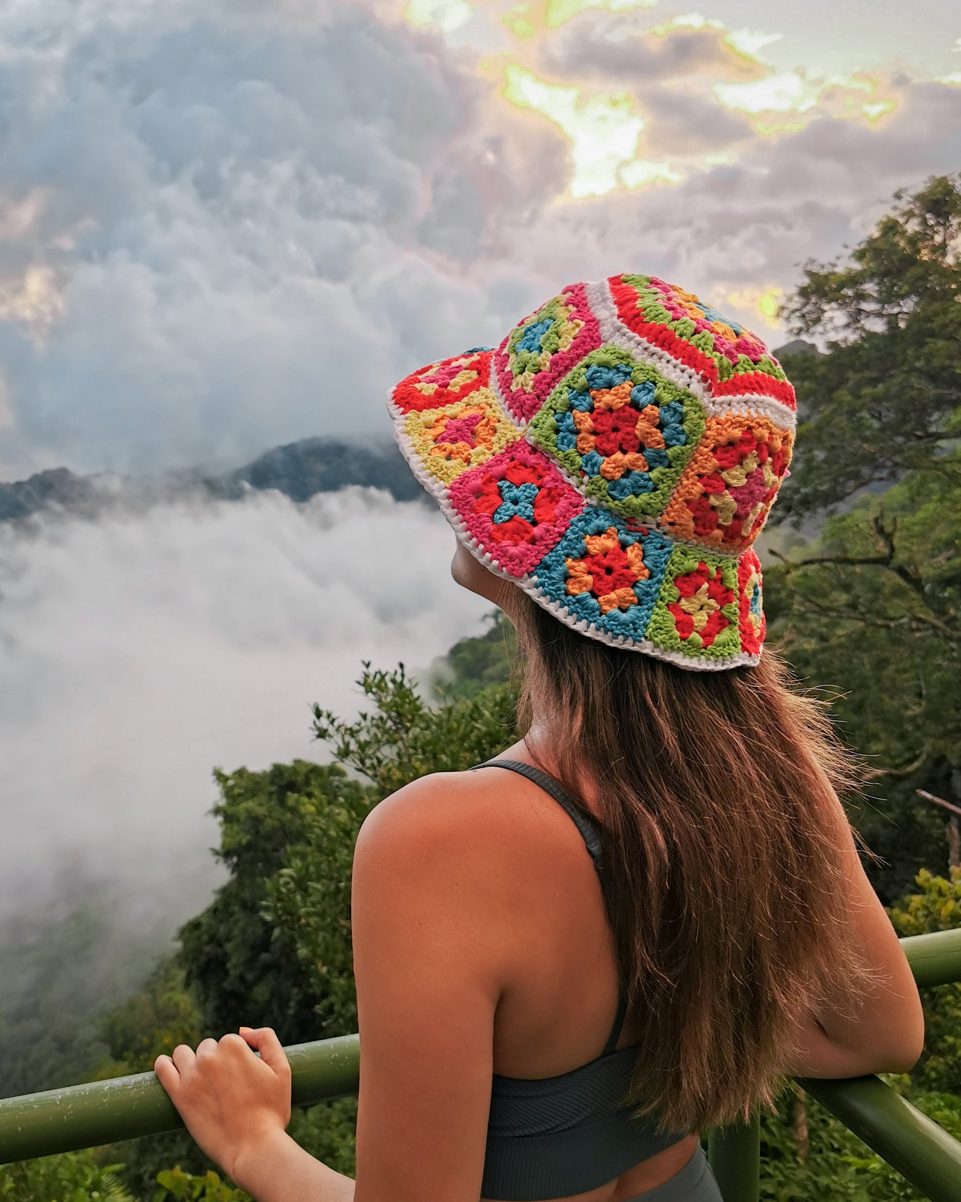 May include: A woman wearing a colorful crochet bucket hat with a white background and a variety of colors including red, orange, yellow, green, blue, and purple. The hat is worn on her head and she is looking out at a view of a mountain range with clouds in the distance.