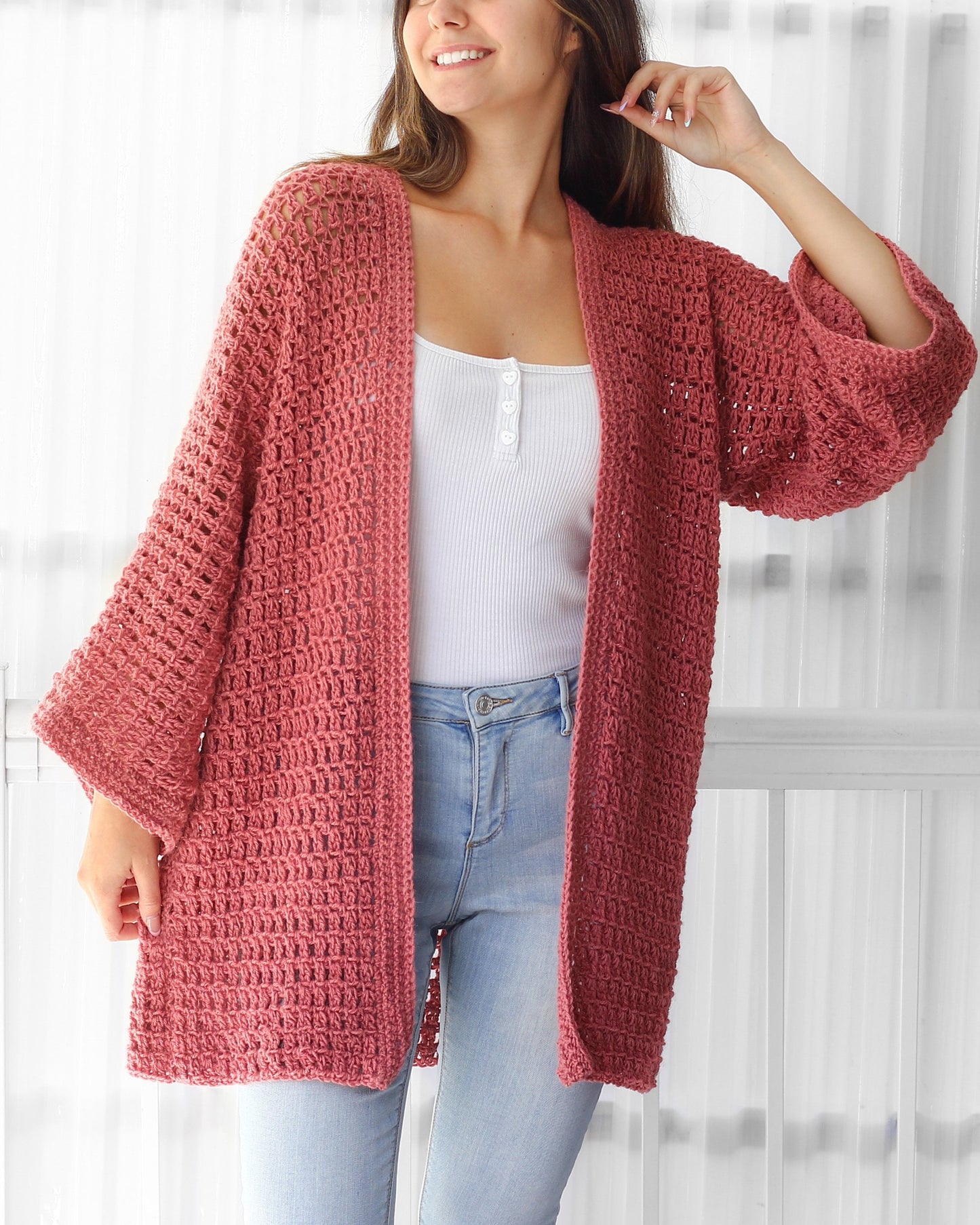 May include: A long, open-front, crocheted cardigan in a dusty rose color. The cardigan has a loose fit and a wide, flowing silhouette. It is made with a delicate, open stitch pattern.
