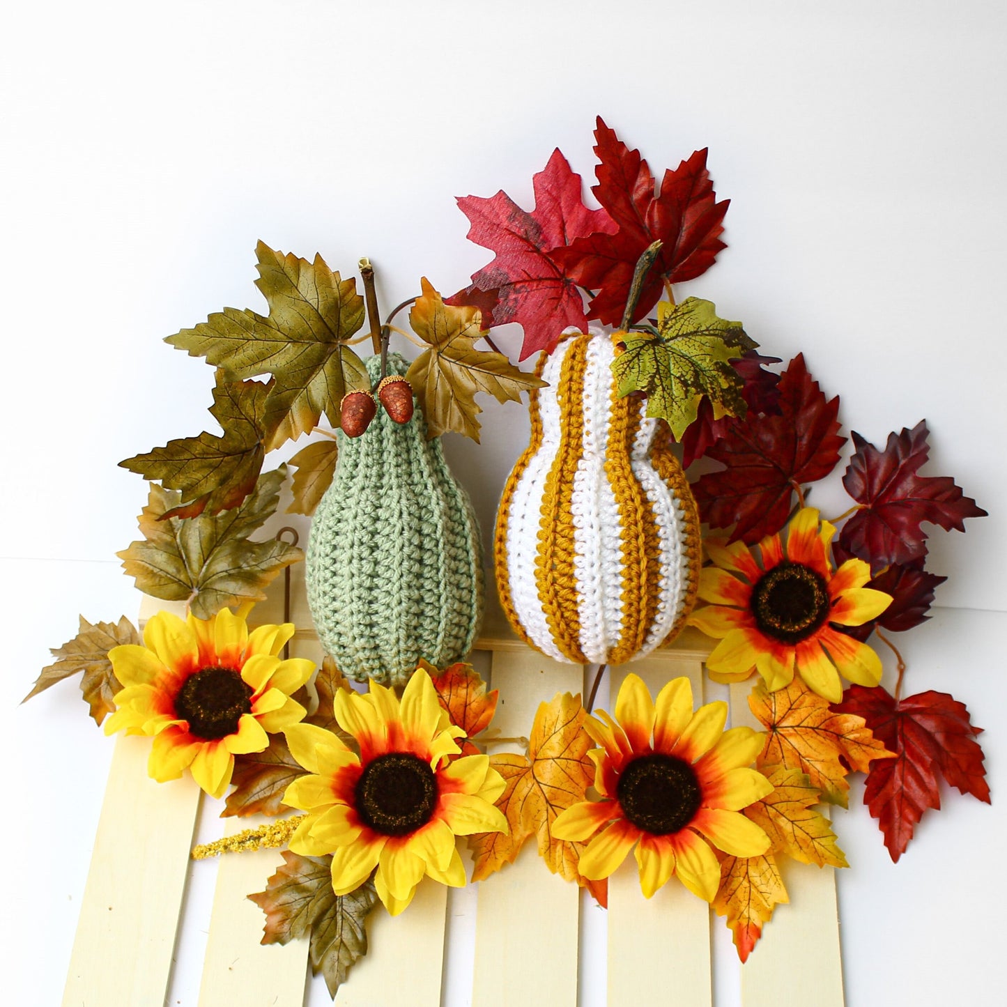 May include: Two crocheted gourds, one green and one striped white and orange, are surrounded by fall foliage, including sunflowers and maple leaves.