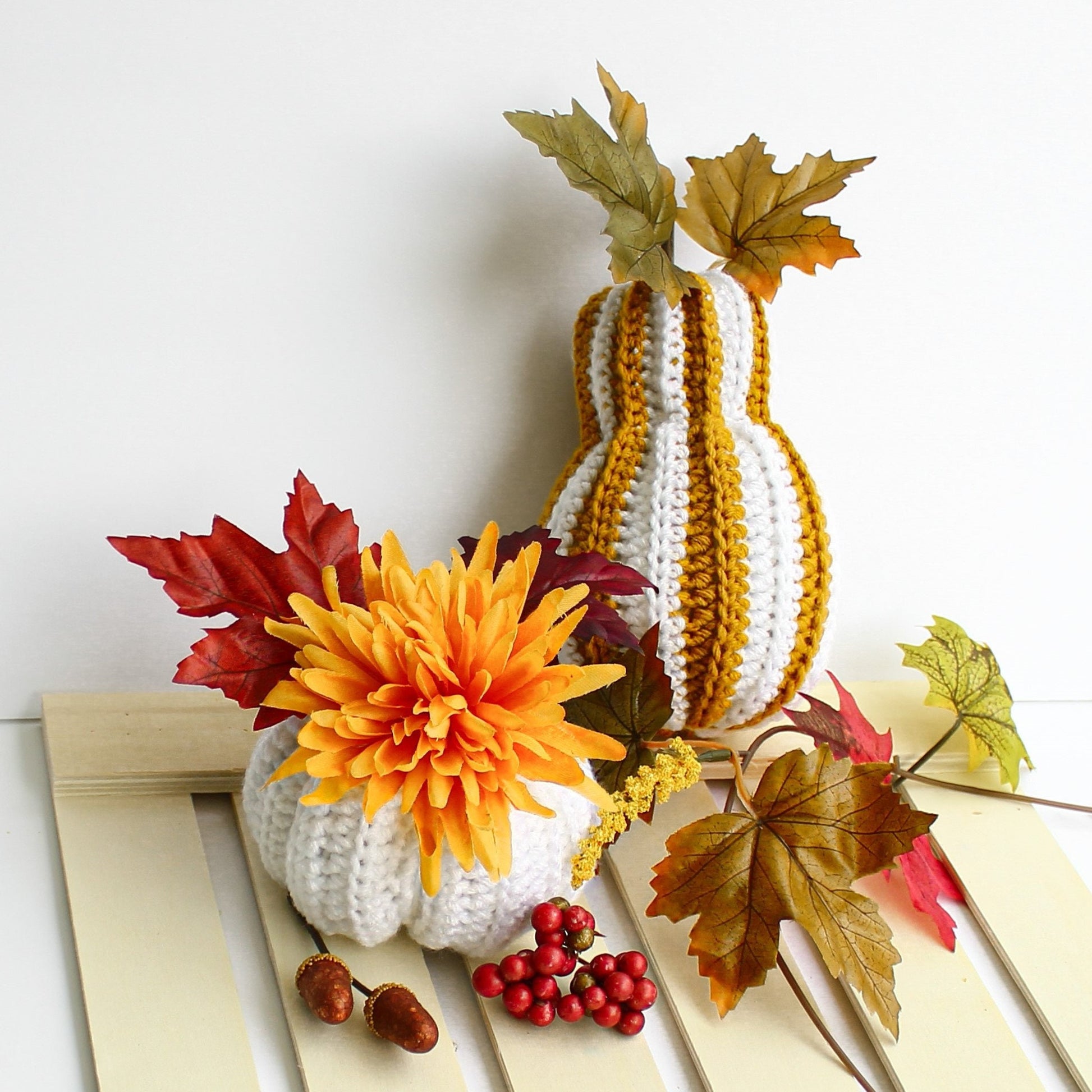 May include: Two crocheted pumpkins decorated with fall foliage. The larger pumpkin is striped with white and yellow yarn. The smaller pumpkin is white with a yellow flower on top.