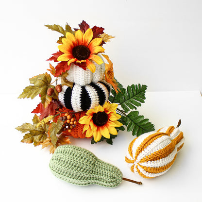 May include: A stack of crocheted pumpkins in white, black and orange with a sunflower on top. The pumpkins are decorated with fall leaves and berries. There are two additional crocheted pumpkins in green and yellow and white stripes.