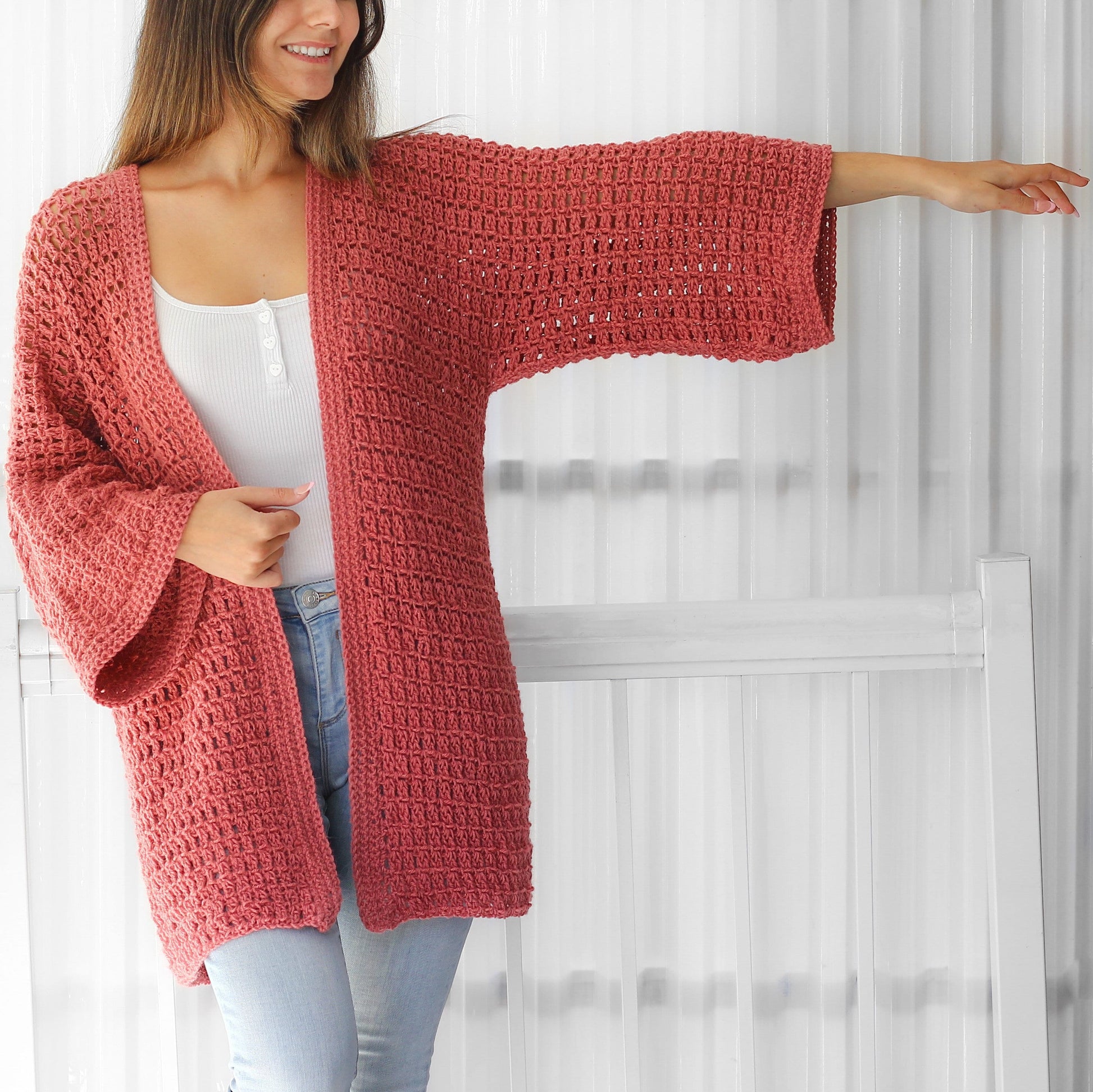 May include: A long, loose-fitting, crocheted cardigan in a dusty rose color. The cardigan has a wide, open front and long sleeves with a bell shape at the cuffs.