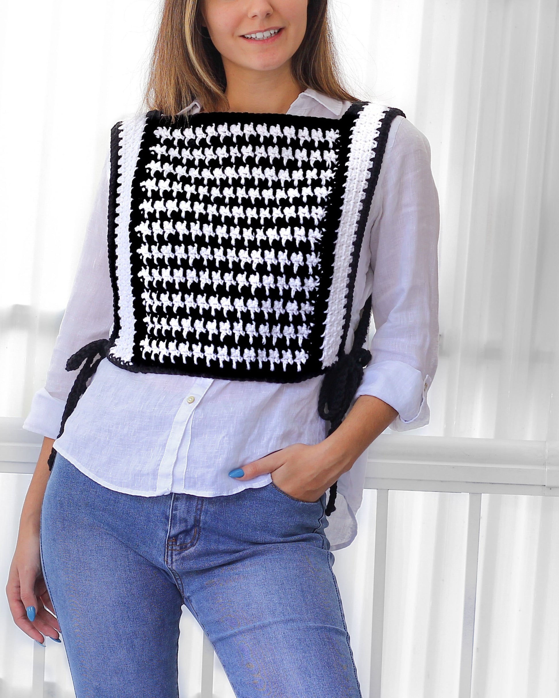 May include: A black and white crocheted vest with a houndstooth pattern. The vest has a square shape and is worn over a white button-down shirt and blue jeans.