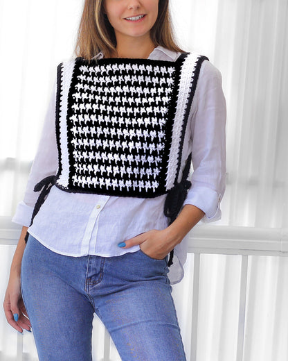 May include: A black and white crocheted vest with a houndstooth pattern. The vest has a square shape and is worn over a white button-down shirt and blue jeans.