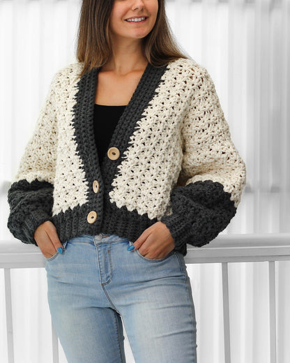 May include: A cropped cardigan with a white and gray color block design. The cardigan has a V-neck and button closure. The sleeves are long and have a slightly puffed look. The cardigan is made of a soft, textured yarn.