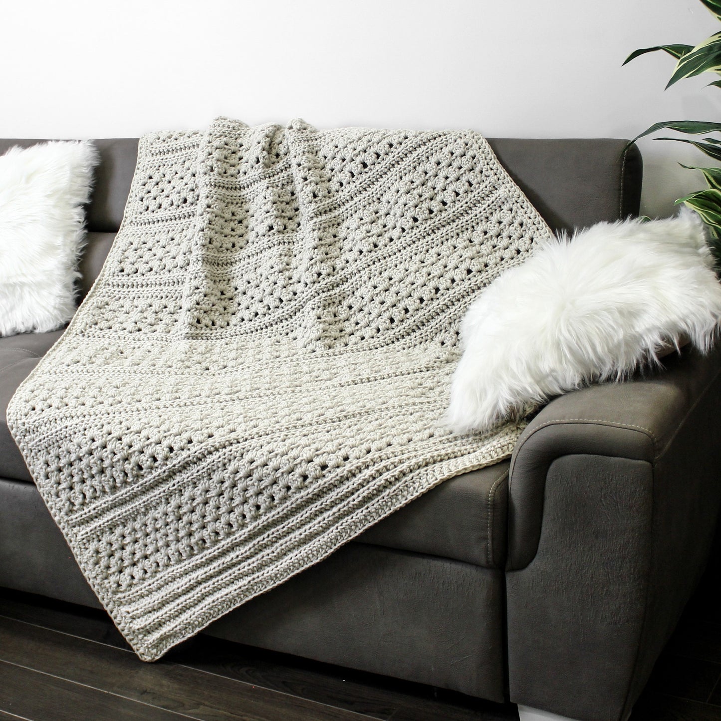 May include: A beige crocheted throw blanket draped over a gray couch with a white faux fur pillow.